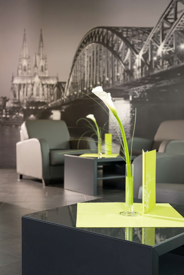 A modern living room with a glass coffee table, a green vase with a white flower, and a framed picture of a bridge in the background. - Essential by Dorint Köln - Junkersdorf (Cologne)