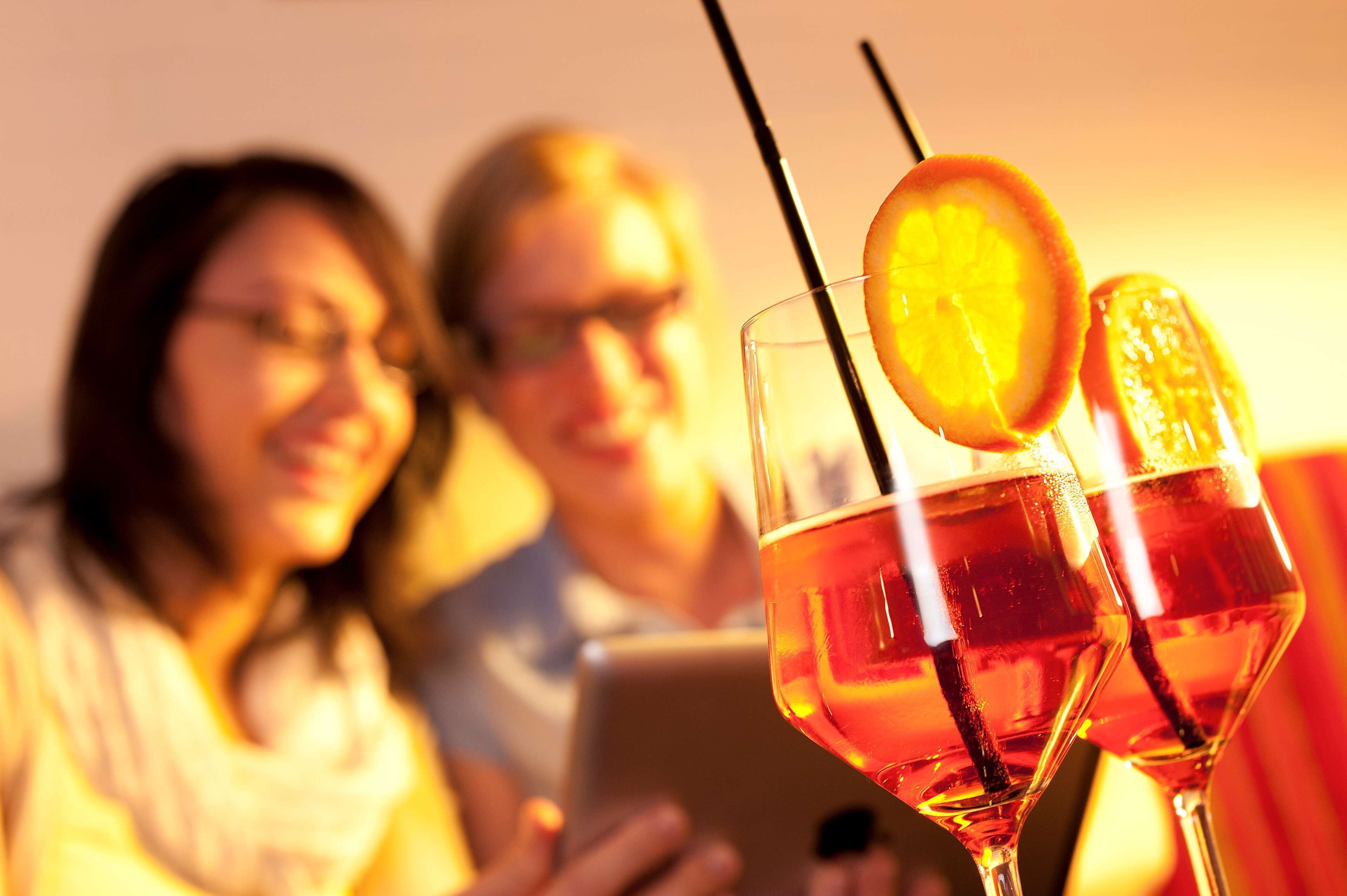 A close-up of two glasses with a drink and a slice of orange, with a blurred background of people.