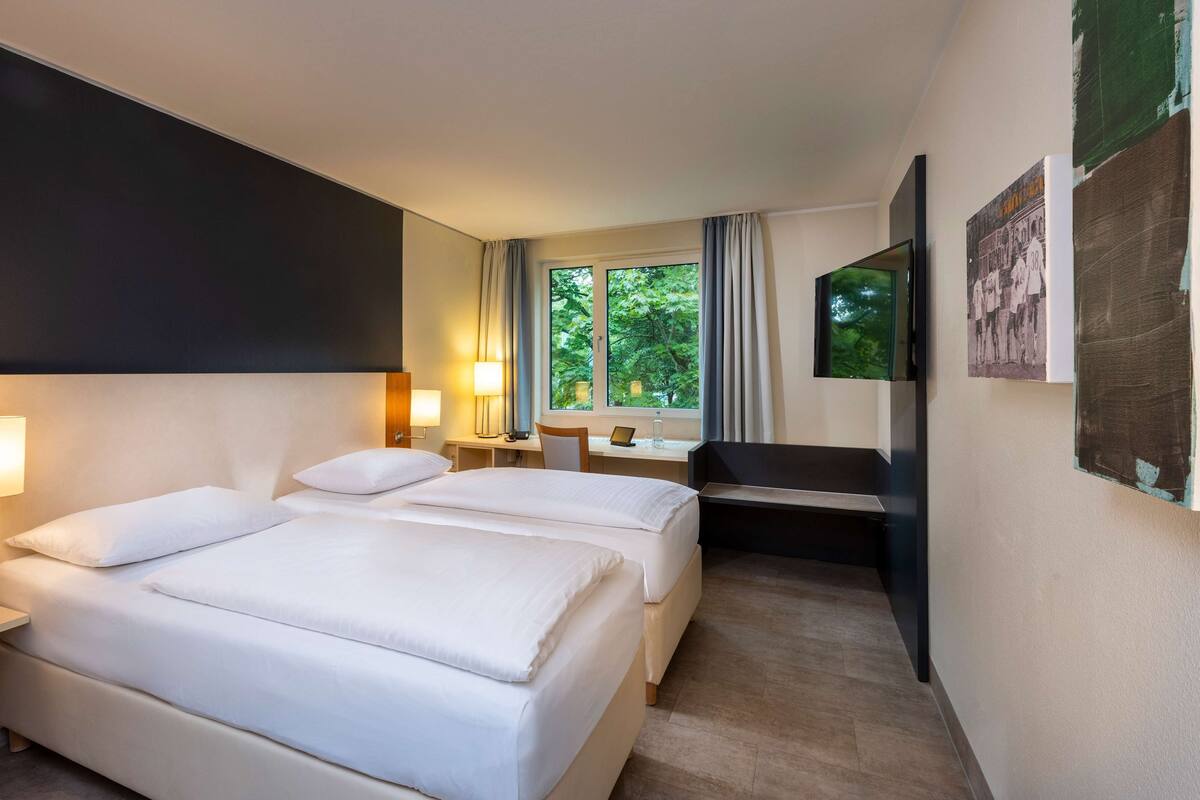 A hotel room with two beds, a desk, a chair, a TV, and a window with a view of trees.
