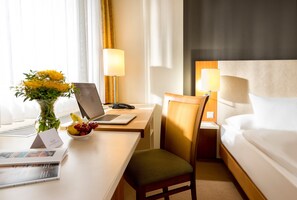 A hotel room with a bed, a desk with a laptop, a chair, a bedside table with a lamp, and a vase with flowers. - Essential by Dorint Köln - Junkersdorf (Cologne)
