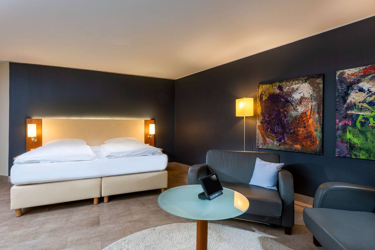 A modern hotel room with a bed, a sofa, a small table with a tablet, and abstract art on the walls.