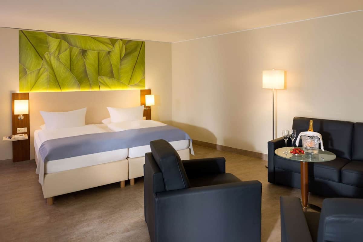 A hotel room with a bed, two bedside tables, a nightstand, a sofa, a small table with a bottle and glasses, and a green leaf-patterned wall.