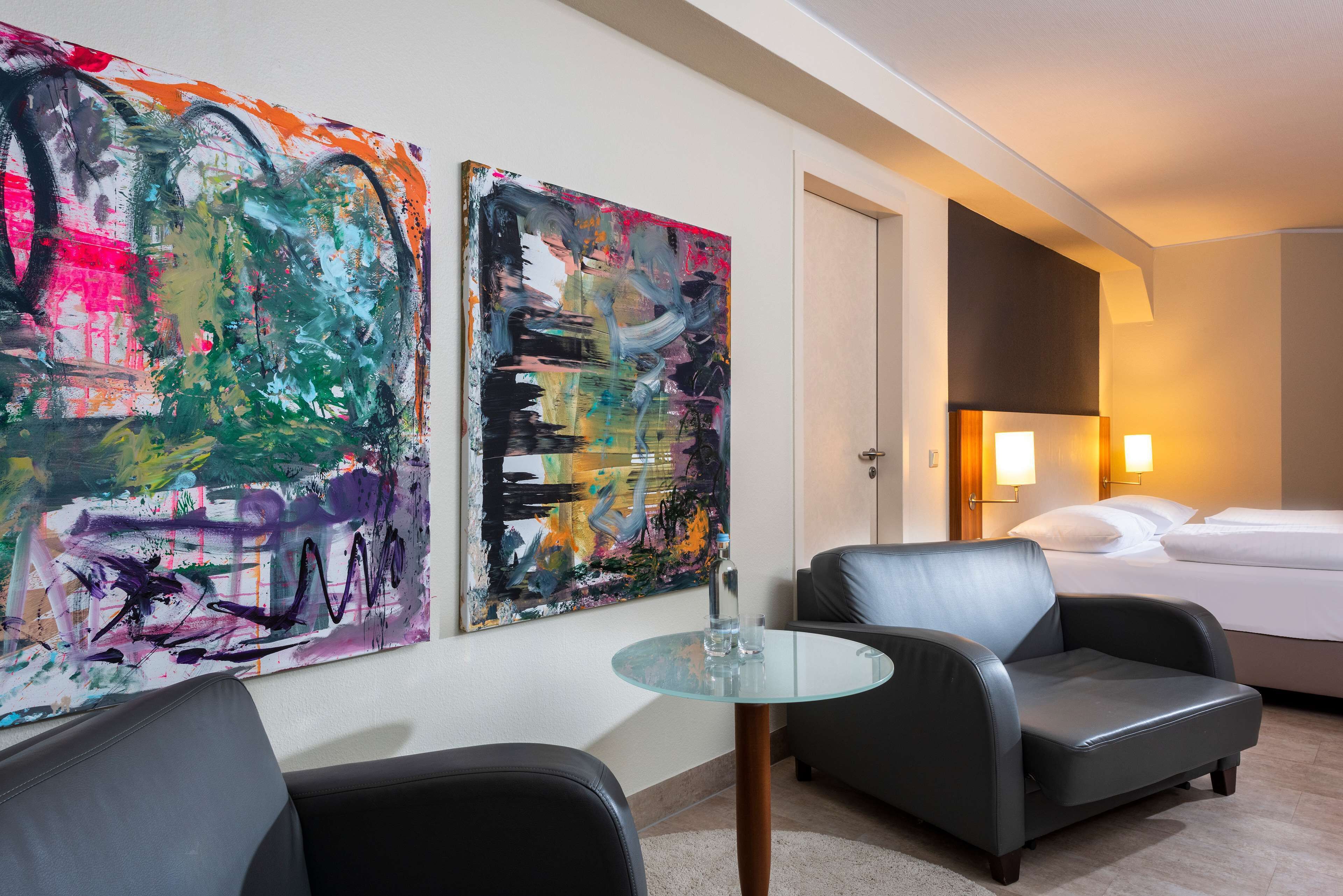 A hotel room with a bed, two armchairs, a glass table, and two abstract paintings on the wall.