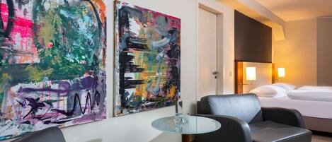 A hotel room with a bed, two armchairs, a glass table, and two abstract paintings on the wall.