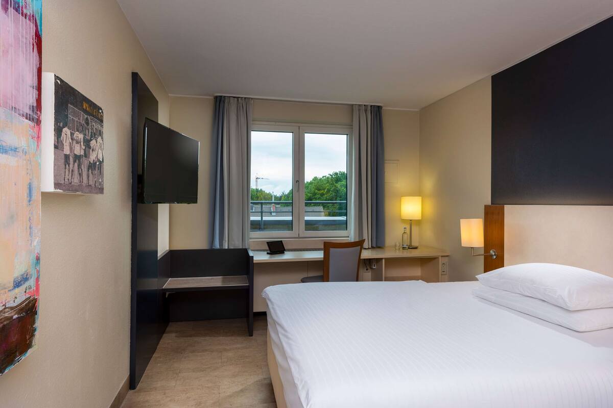 A hotel room with a bed, desk, chair, TV, and a window with curtains.