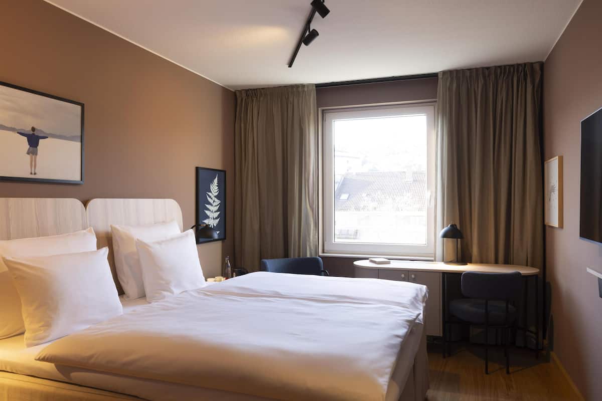 A hotel room with a large bed, a desk, a chair, a window with curtains, and a framed picture on the wall.