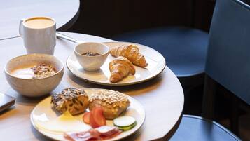 Une table mise pour le petit-déjeuner avec des croissants, un bol de céréales, une tasse de café et une assiette contenant des œufs, des tomates et des concombres.