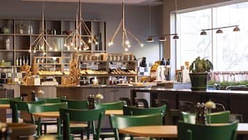 A restaurant interior with green chairs, wooden tables, hanging pendant lights, shelves with bottles and food items, and a large window with a view.