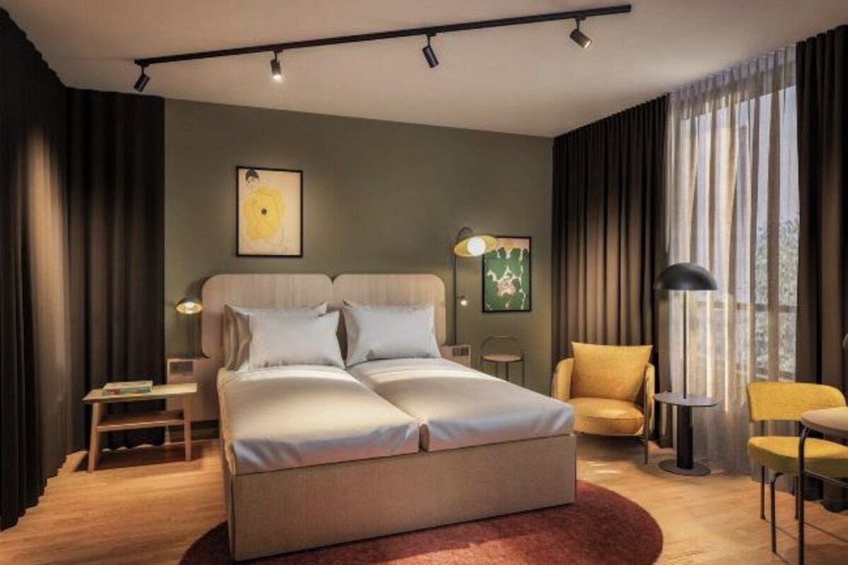A modern hotel room with a large bed, a desk, a chair, and a lamp.