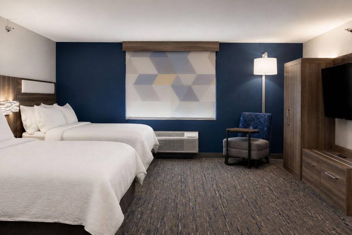 A hotel room with two beds, a desk, a chair, a TV, and a window with a geometric pattern.