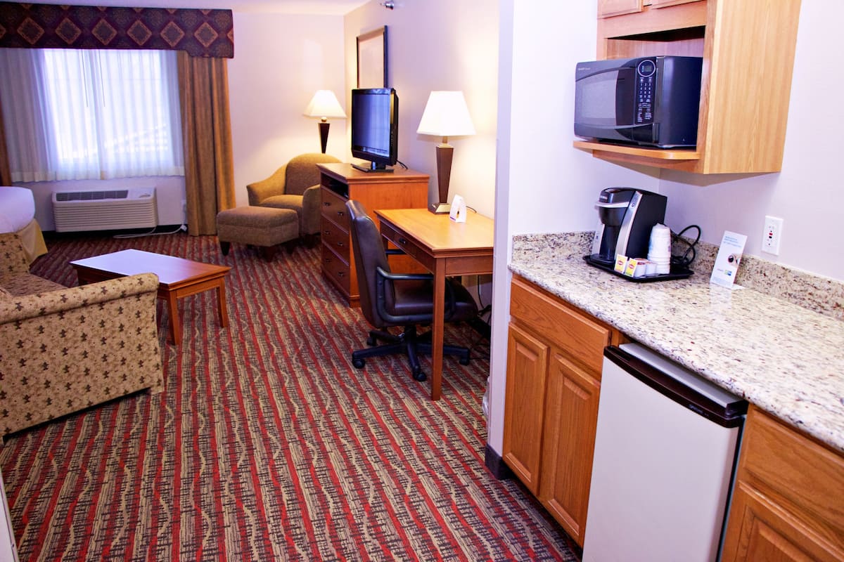 A hotel room with a kitchenette, a sofa, a desk, a chair, a television, a microwave, and a coffee maker.