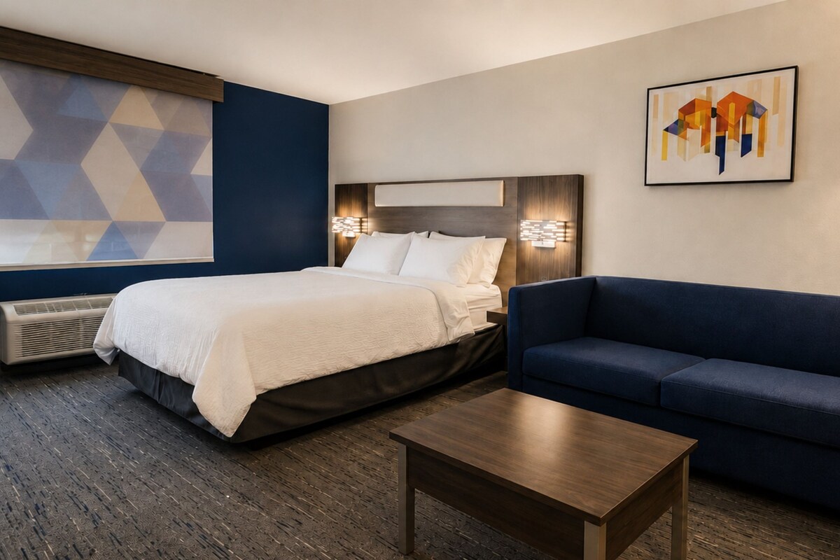 A hotel room with a bed, a blue sofa, a wooden coffee table, a wall-mounted lamp, and a geometric-patterned wall.
