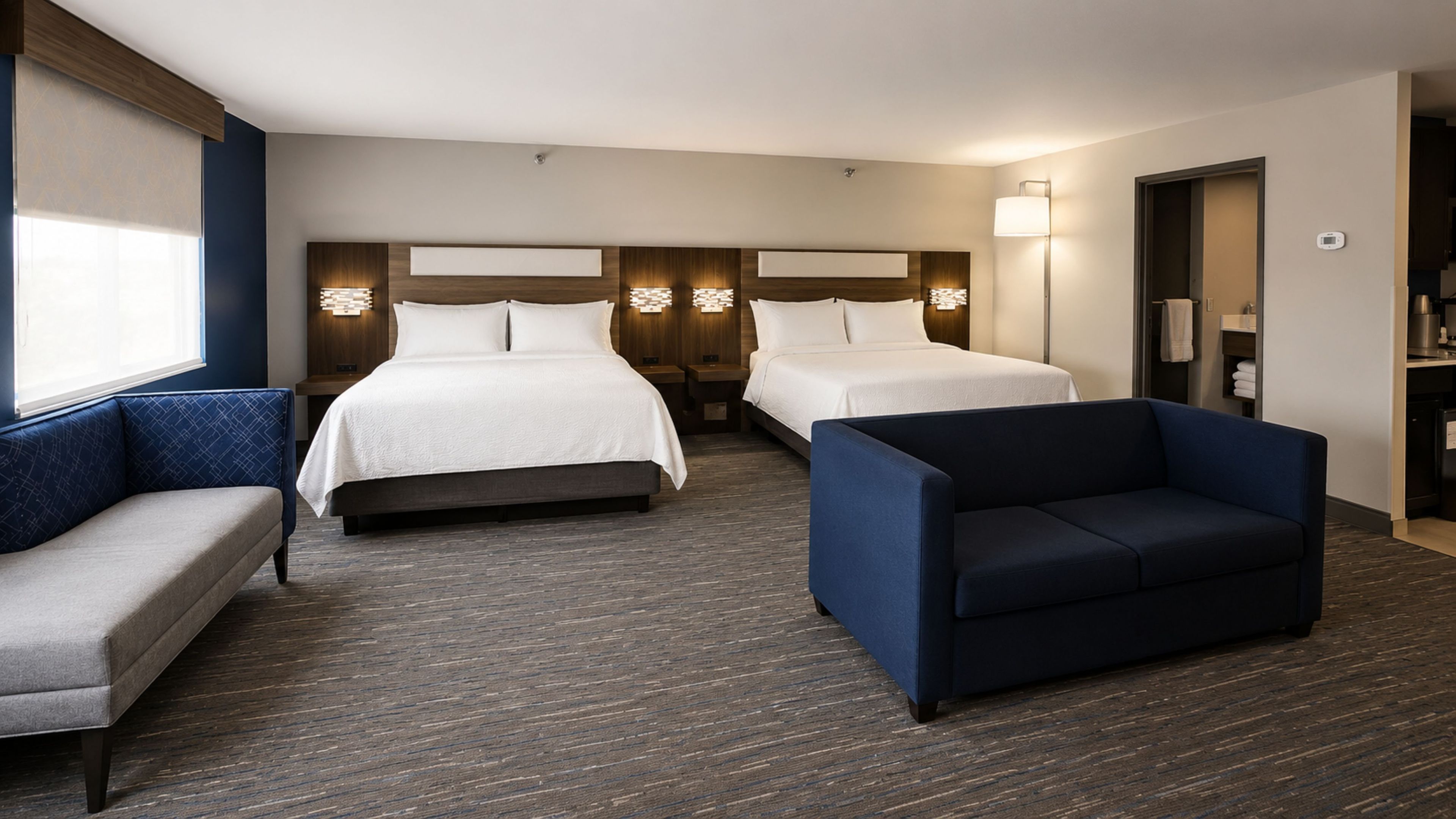 Quarto de hotel com duas camas, um sofá, uma poltrona, uma mesa de cabeceira, um abajur e uma pequena cozinha equipada.