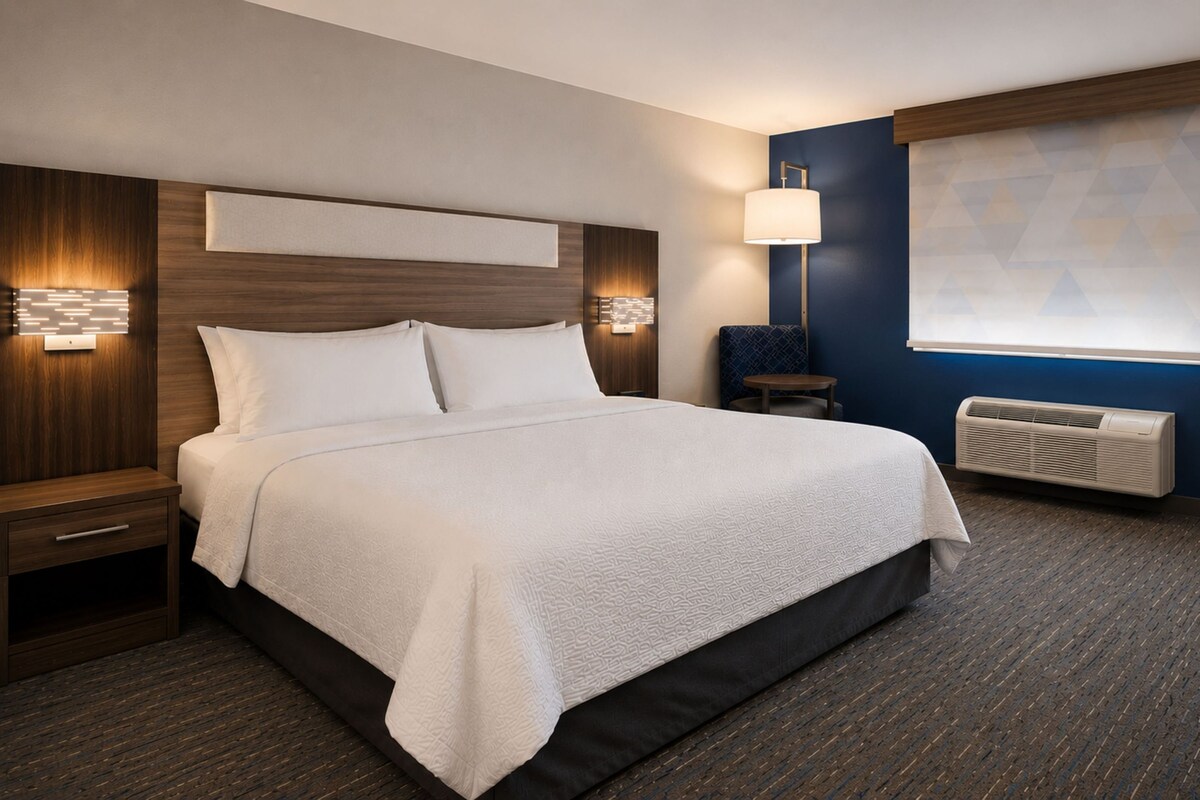 A hotel room with a large bed, bedside tables, a lamp, a window with blinds, and a wall-mounted air conditioner.