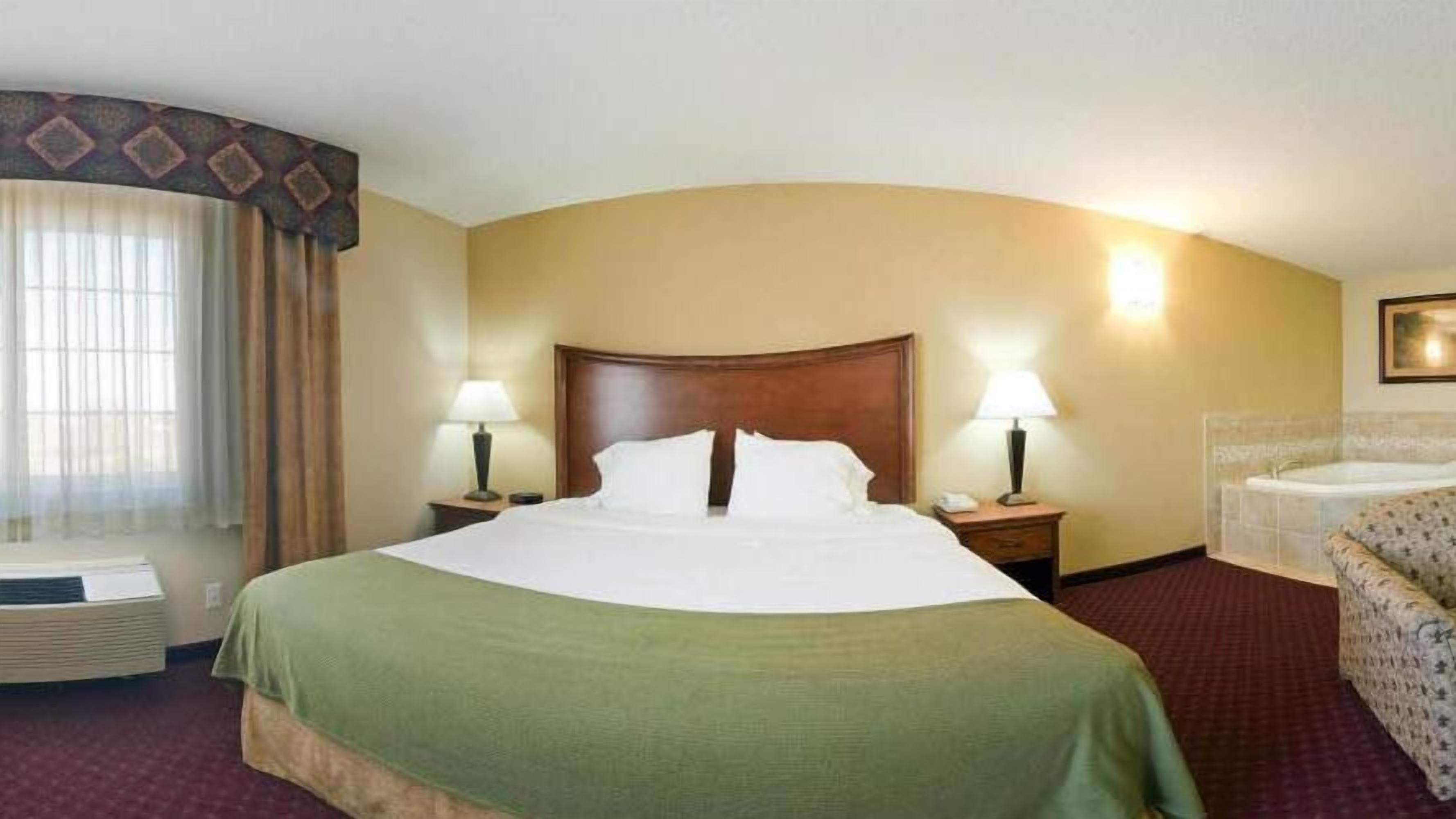 A hotel room with a large bed, two bedside tables with lamps, a sofa, a window with curtains, and a wall-mounted light fixture.