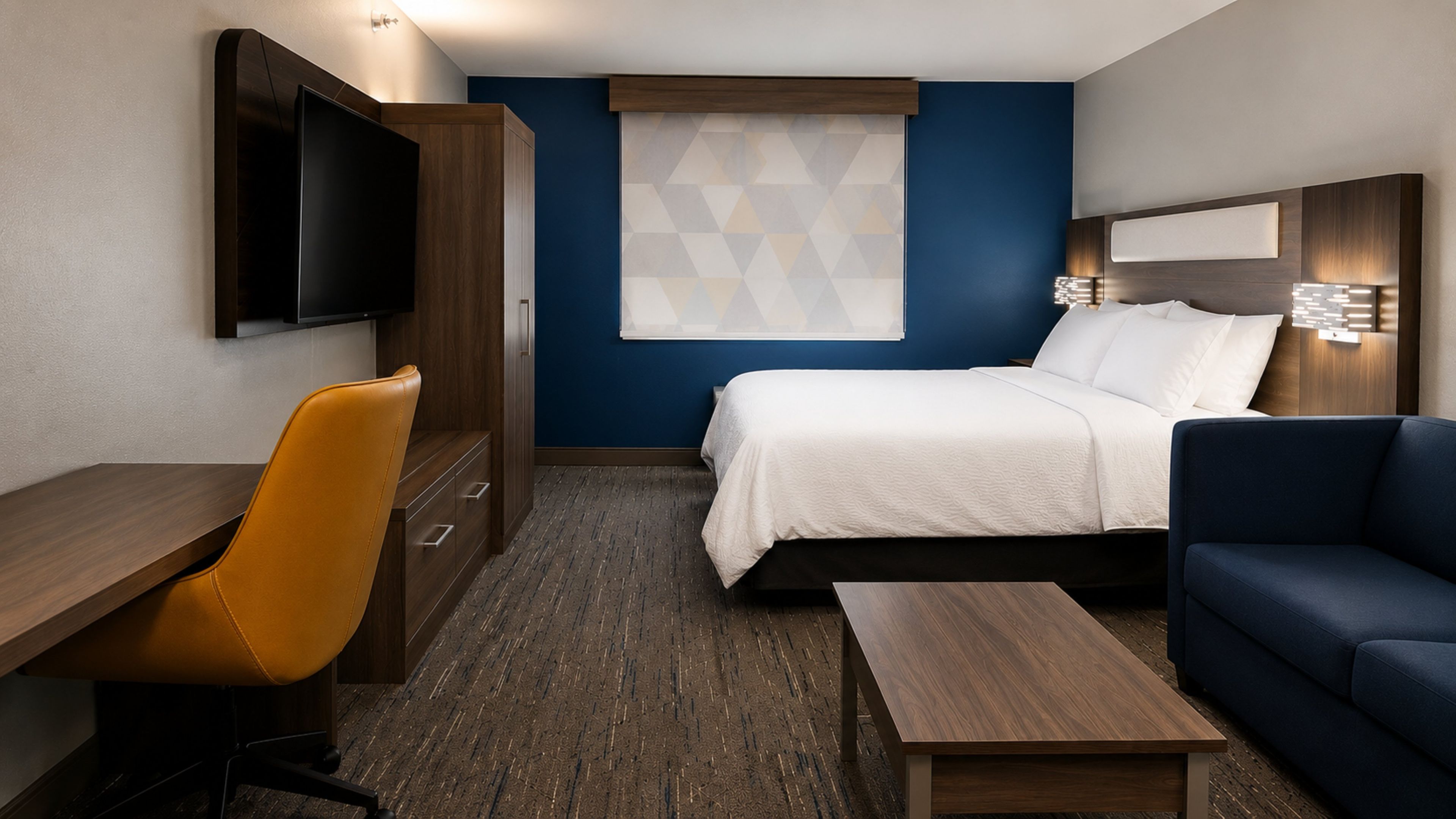 Ein Hotelzimmer mit einem Bett, einem Schreibtisch, einem Sessel, einer Couch, einem Fernseher und einem Fenster mit geometrischem Muster.