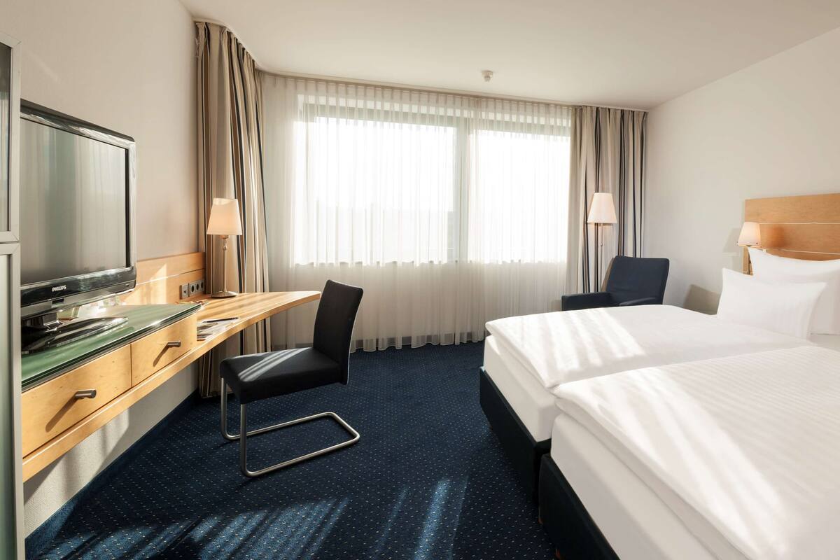 Una habitación de hotel con cama, escritorio, silla, televisor y ventana con cortinas.