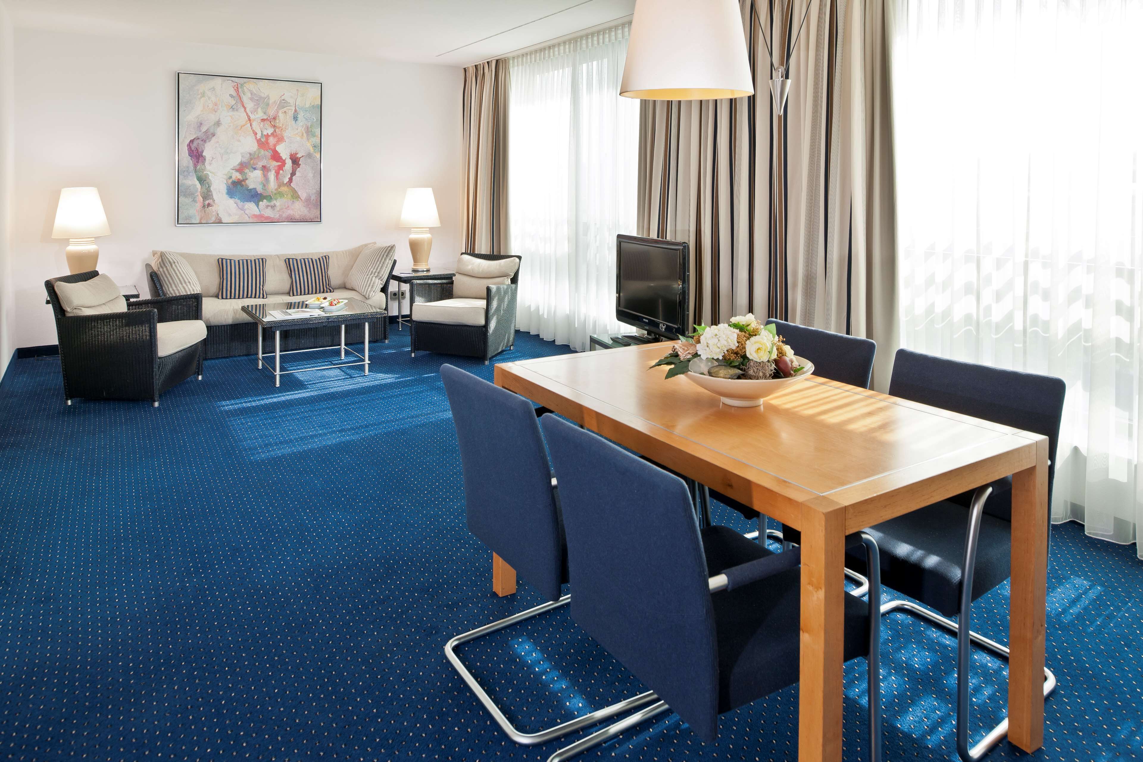 Une chambre d’hôtel avec un tapis bleu, une table à manger avec des chaises, un ensemble de canapés, un téléviseur et une grande fenêtre avec des rideaux.