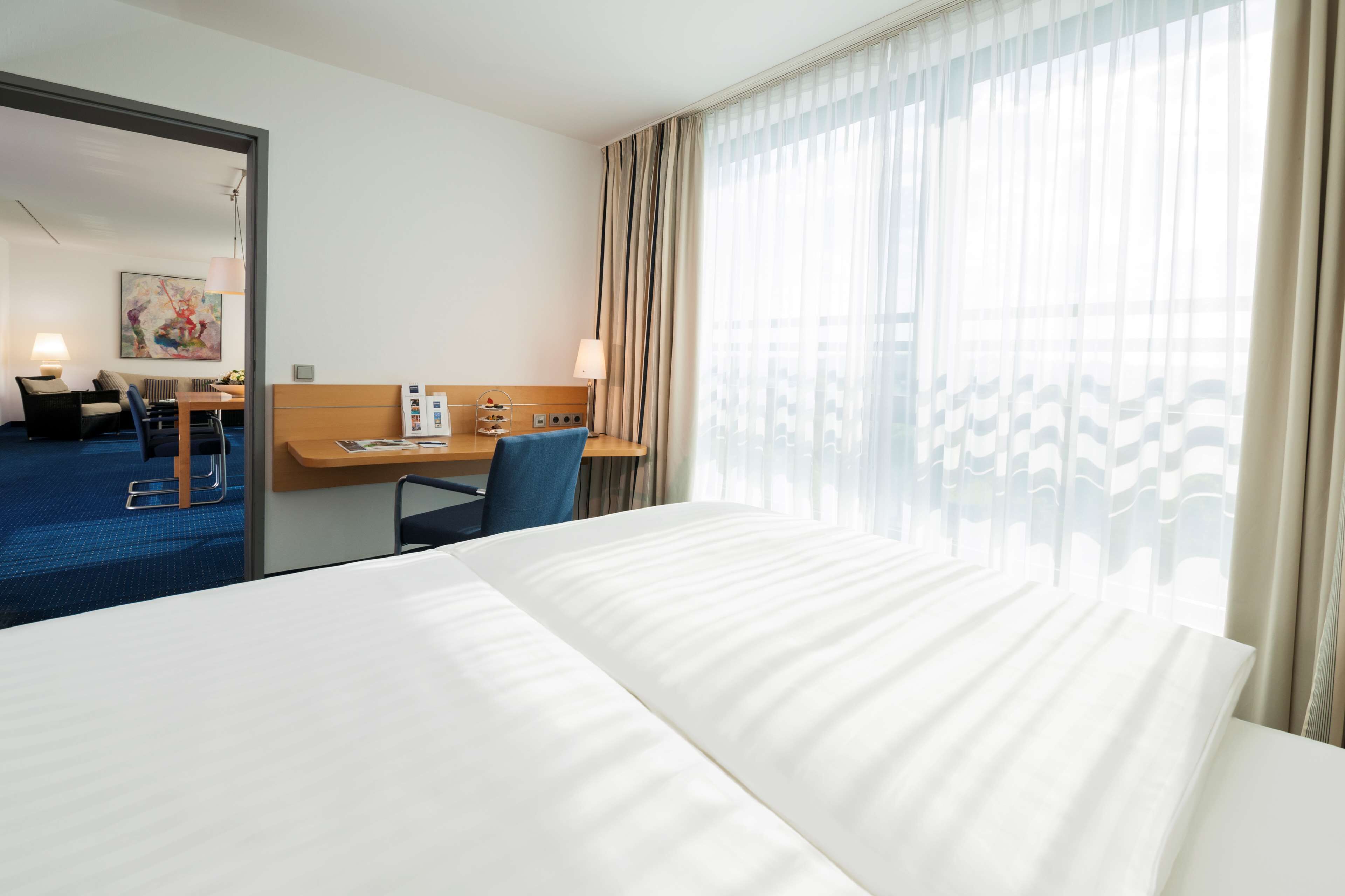 Quarto de hotel com uma cama grande, uma escrivaninha com cadeira, uma mesinha e uma janela com cortinas.