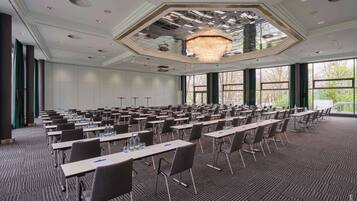 Een ruime congrescentrum met rijen tafels en stoelen, grote ramen en een kroonluchter.