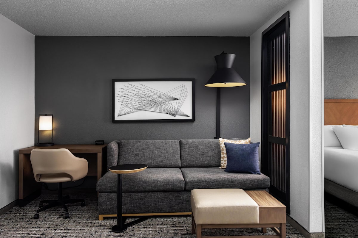 Una habitación moderna con un sofá gris, una pequeña mesa redonda, un escritorio con una silla, una lámpara de pie y una obra de arte abstracta enmarcada en la pared.