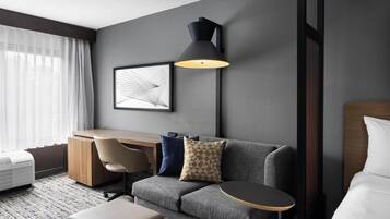 Quarto de hotel moderno com sofá cinzento, uma pequena mesa redonda, uma escrivaninha, uma cadeira, uma luminária grande fixada na parede e um quadro emoldurado.