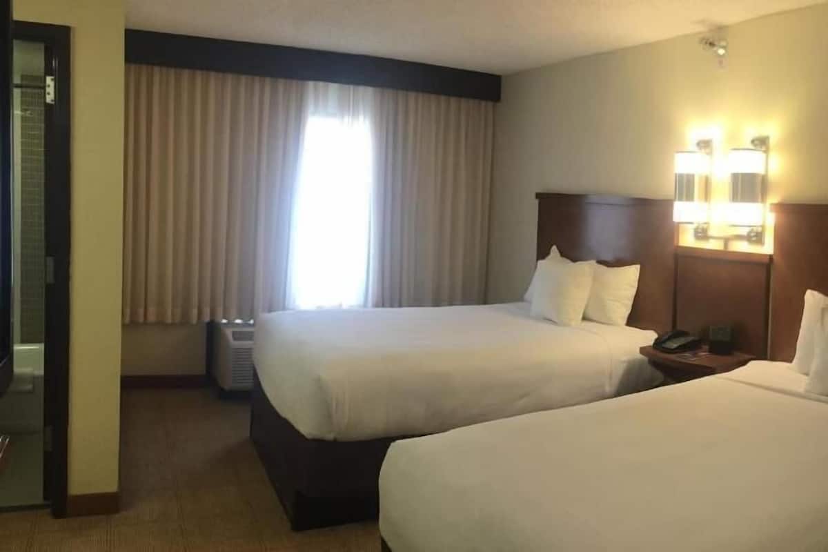 Habitación de hotel con dos camas, una mesita de noche, una lámpara y una ventana con cortinas.