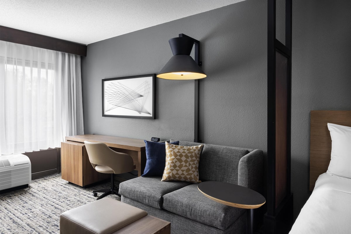 A modern hotel room with a grey sofa, a wooden desk, a black lamp, and a framed picture on the wall.