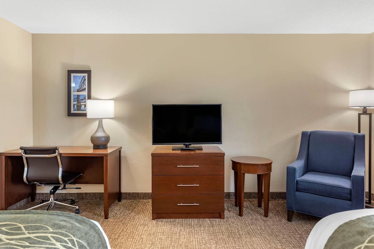 A hotel room with a bed, a desk with a chair, a dresser, a television, a lamp, and a blue armchair.