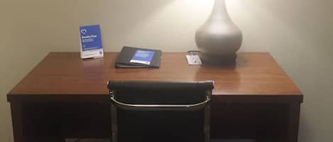 A hotel room with a wooden desk, a black office chair, a framed picture of a bridge, a lamp, and a card on the desk.