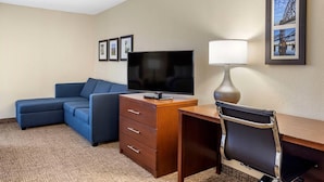 A hotel room with a sectional sofa, a desk with a chair, a flat-screen TV, a lamp, and framed pictures on the wall. - Comfort Inn & Suites Little Rock Airport (Little Rock)