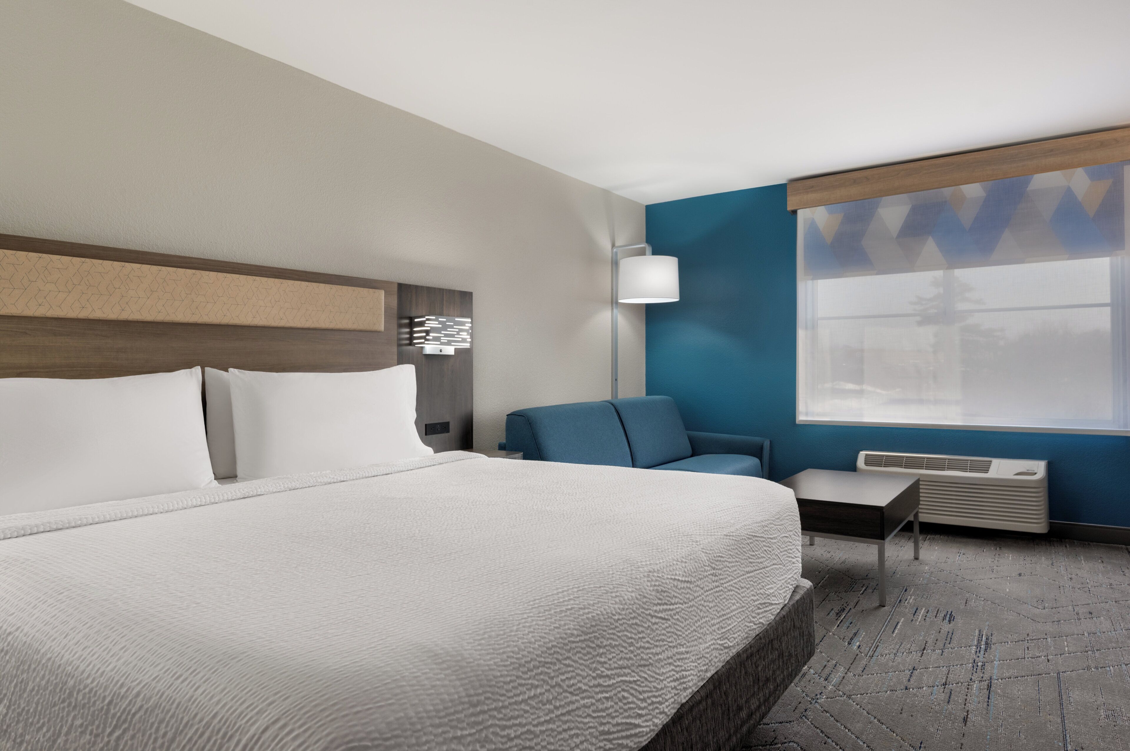 A hotel room with a large bed, a seating area, a window with blinds, and a wall-mounted air conditioner.