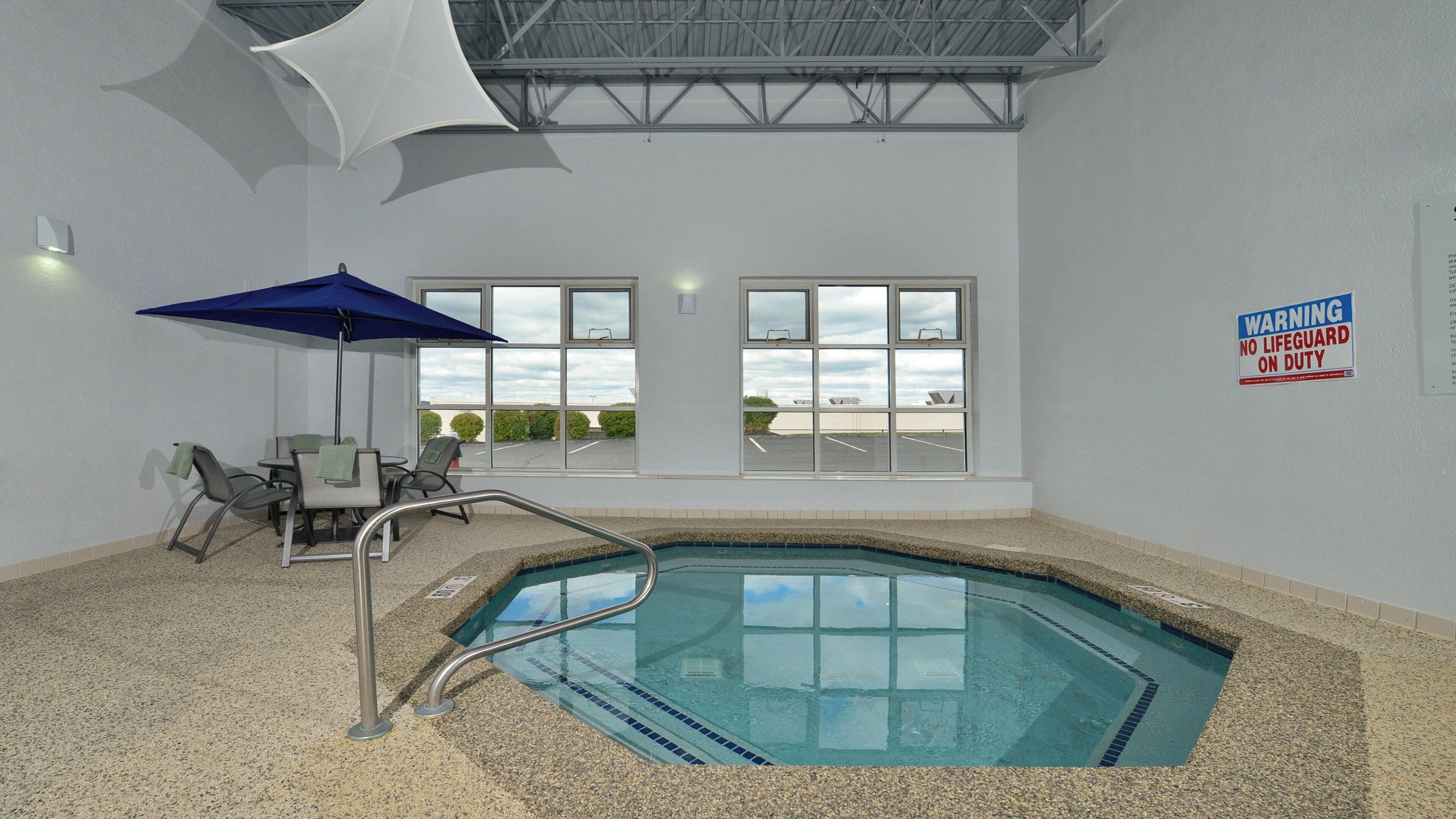 An indoor swimming pool with a lifeguard warning sign, a lounge area with chairs and a table, and large windows offering a view of the parking lot.