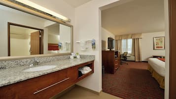 A hotel room with a large mirror, a sink, a wooden vanity, a bed, a desk, and a television.