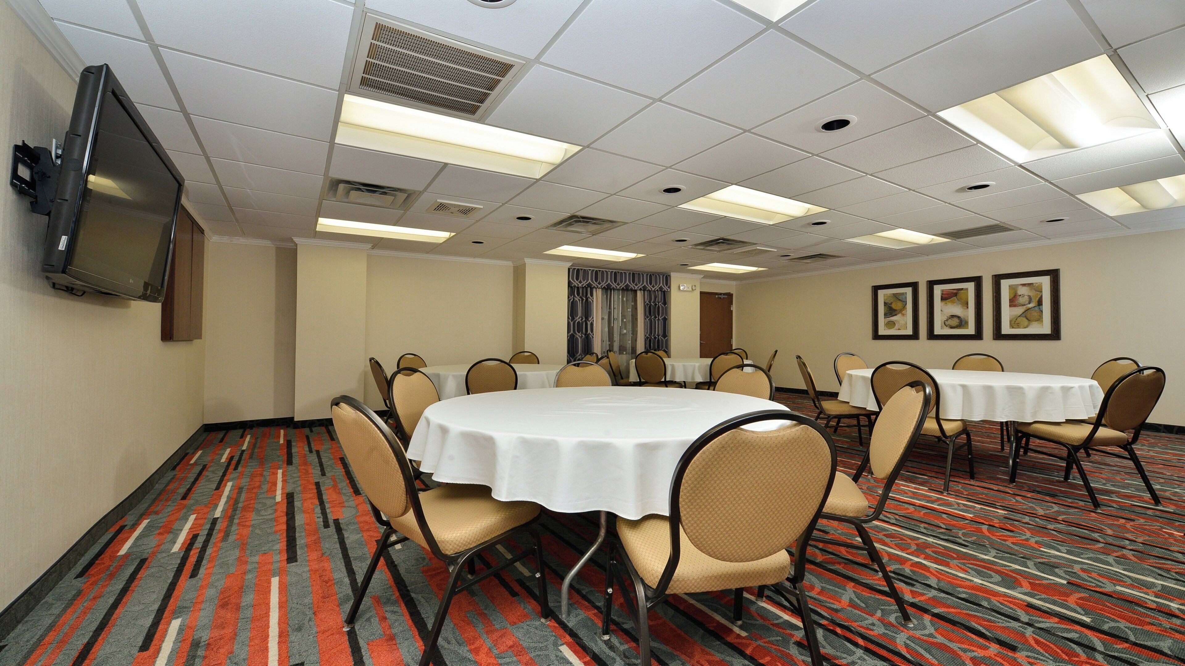 A conference room with multiple tables and chairs, a flat-screen TV mounted on the wall, and patterned carpet flooring.