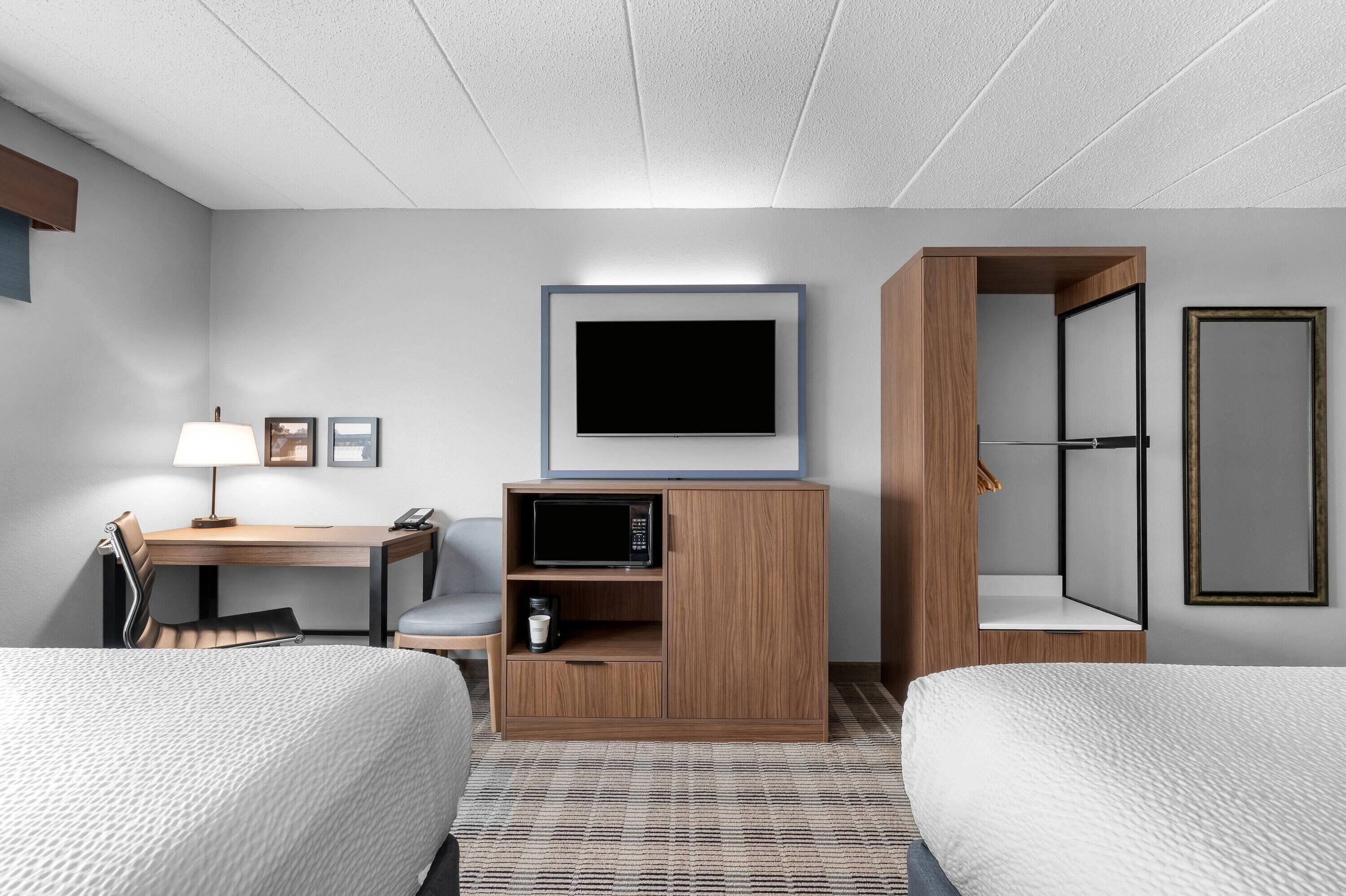 Ein Hotelzimmer mit zwei Betten, einem Schreibtisch mit Lampe, einem Fernseher, einem Kleiderschrank und einer Kaffeemaschine.