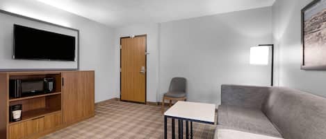 A hotel room with a flat-screen TV, a microwave, a wooden cabinet, a gray sofa, a small table, and a door.