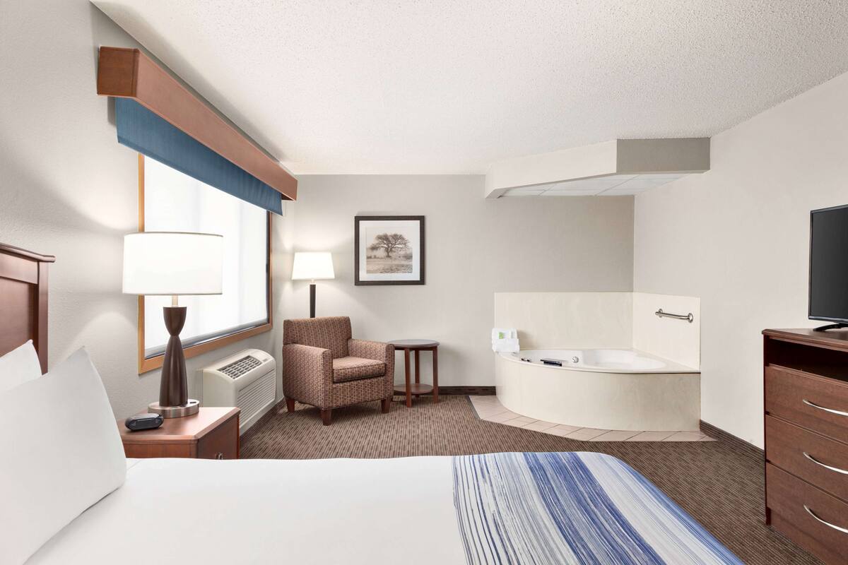 A hotel room with a bed, a chair, a small table, a lamp, a picture on the wall, a bathtub, and a TV.