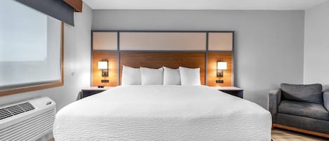 Ein Hotelzimmer mit einem großen Bett, einem hölzernen Kopfteil, zwei Nachttischlampen, einem grauen Sofa und einem Fenster mit Jalousien.