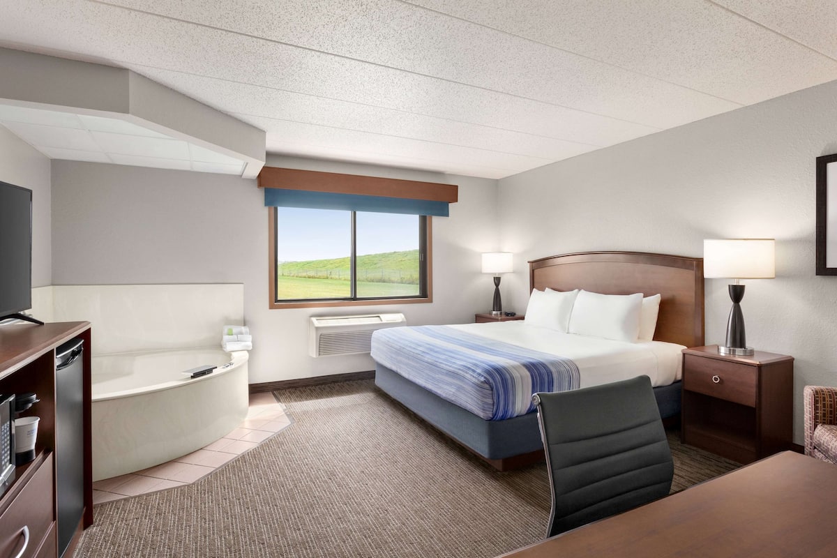 A hotel room with a large bed, a desk, a chair, a desk lamp, a bedside table, a lamp, a TV, a bathroom with a bathtub, and a window with a view of a green landscape.
