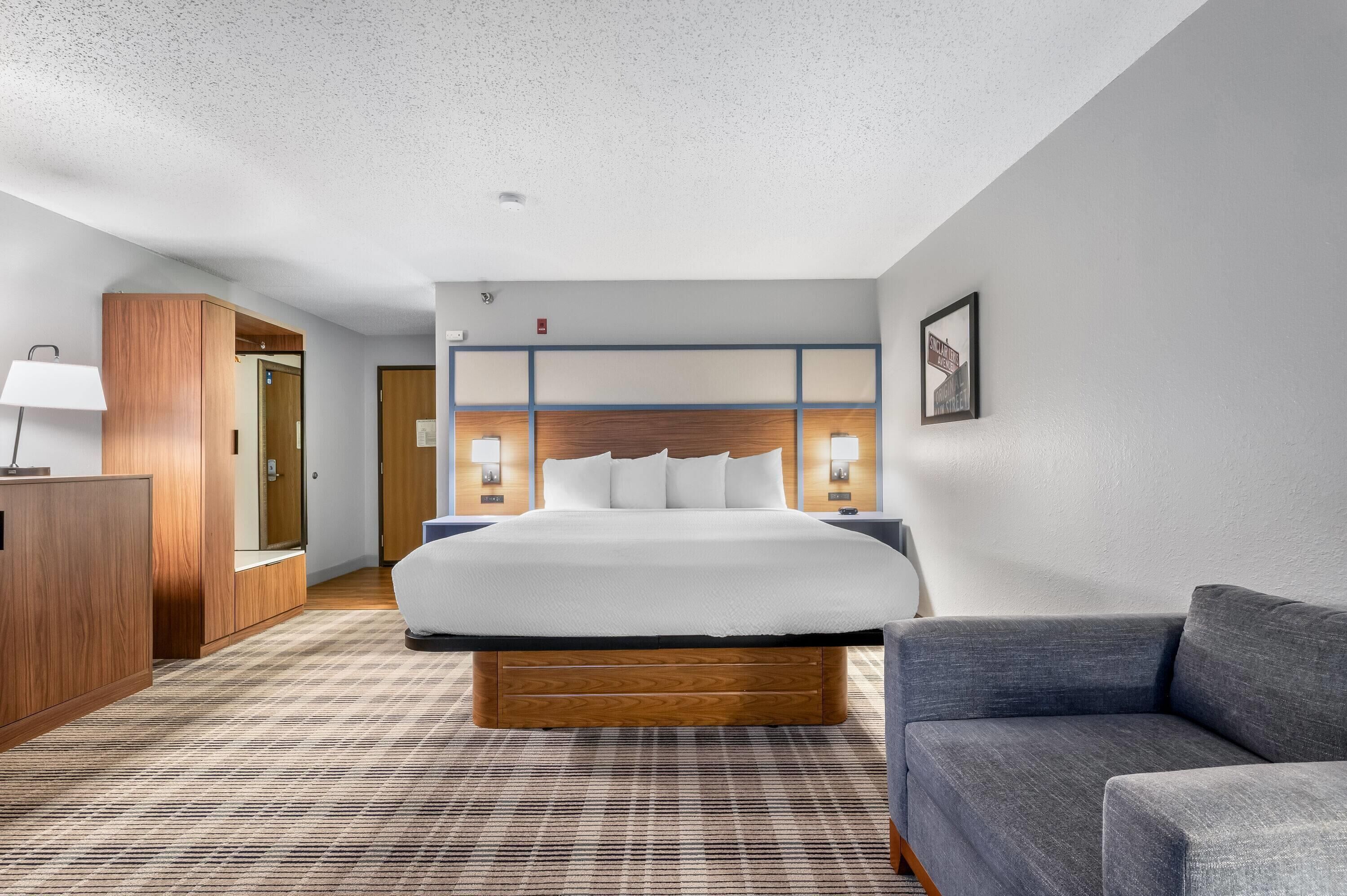 A hotel room with a large bed, a wooden headboard, a nightstand, a desk, a lamp, a couch, and a patterned carpet.