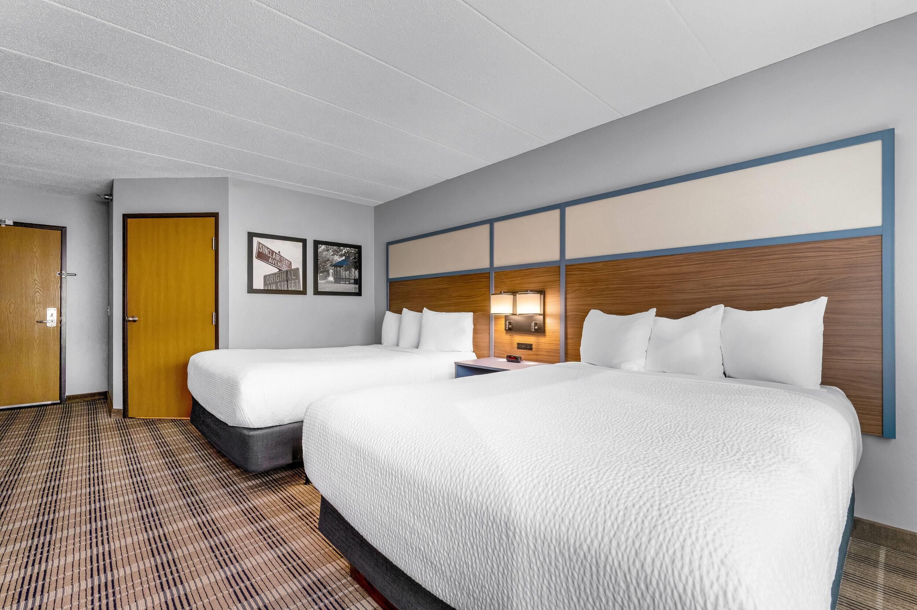 A hotel room with two beds, a wooden door, a wall-mounted lamp, and a check-in desk.
