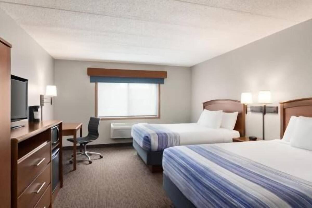 Une chambre d’hôtel avec deux lits, un bureau avec une chaise, une télévision et une fenêtre avec des stores.