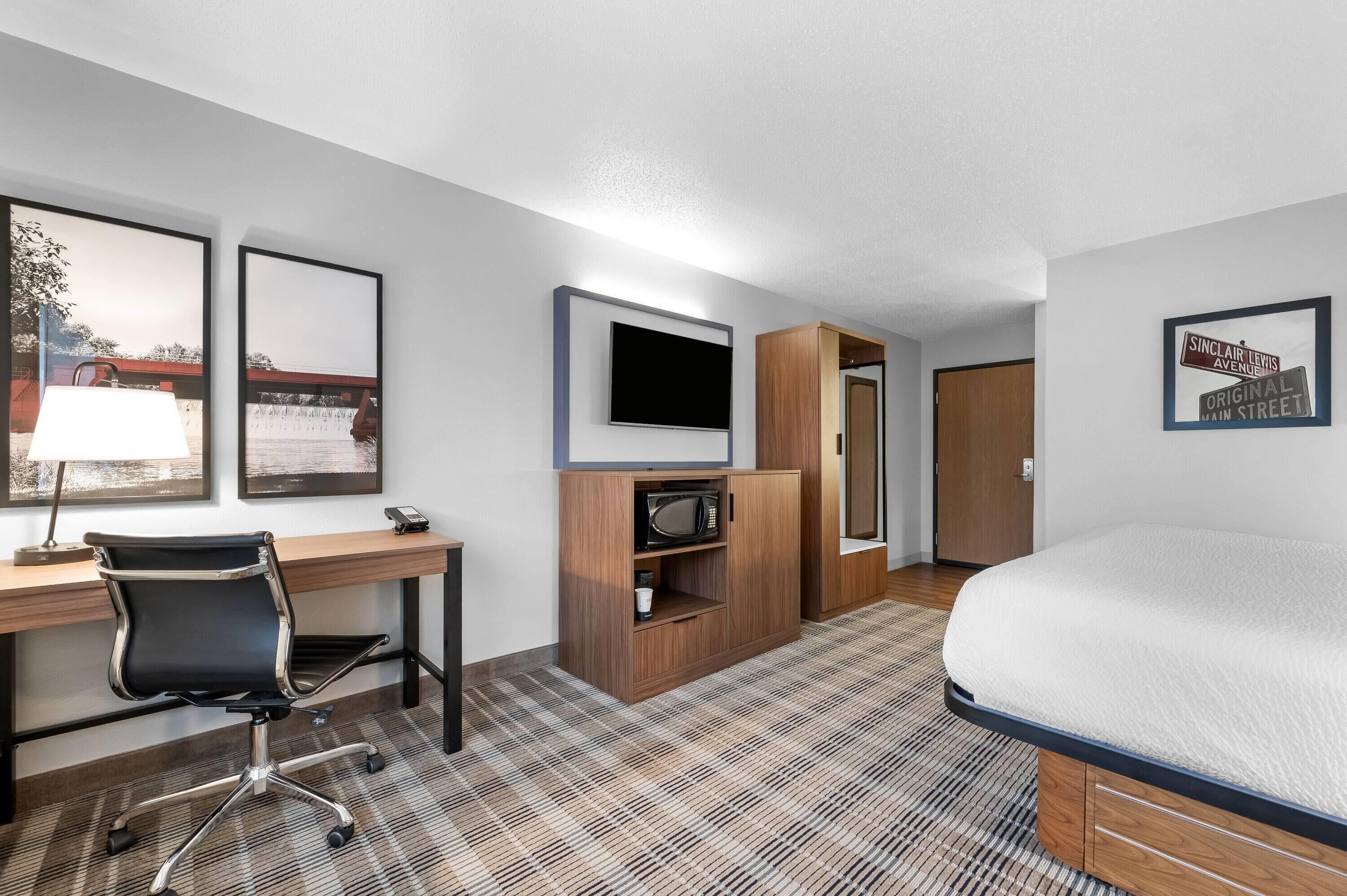 A hotel room with a bed, desk, chair, television, and framed pictures on the wall.