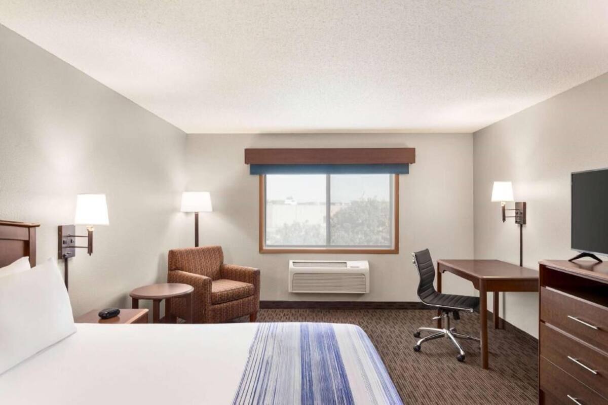 Une chambre d’hôtel comprenant un lit, deux chaises, un bureau, une télévision et une fenêtre avec des stores.