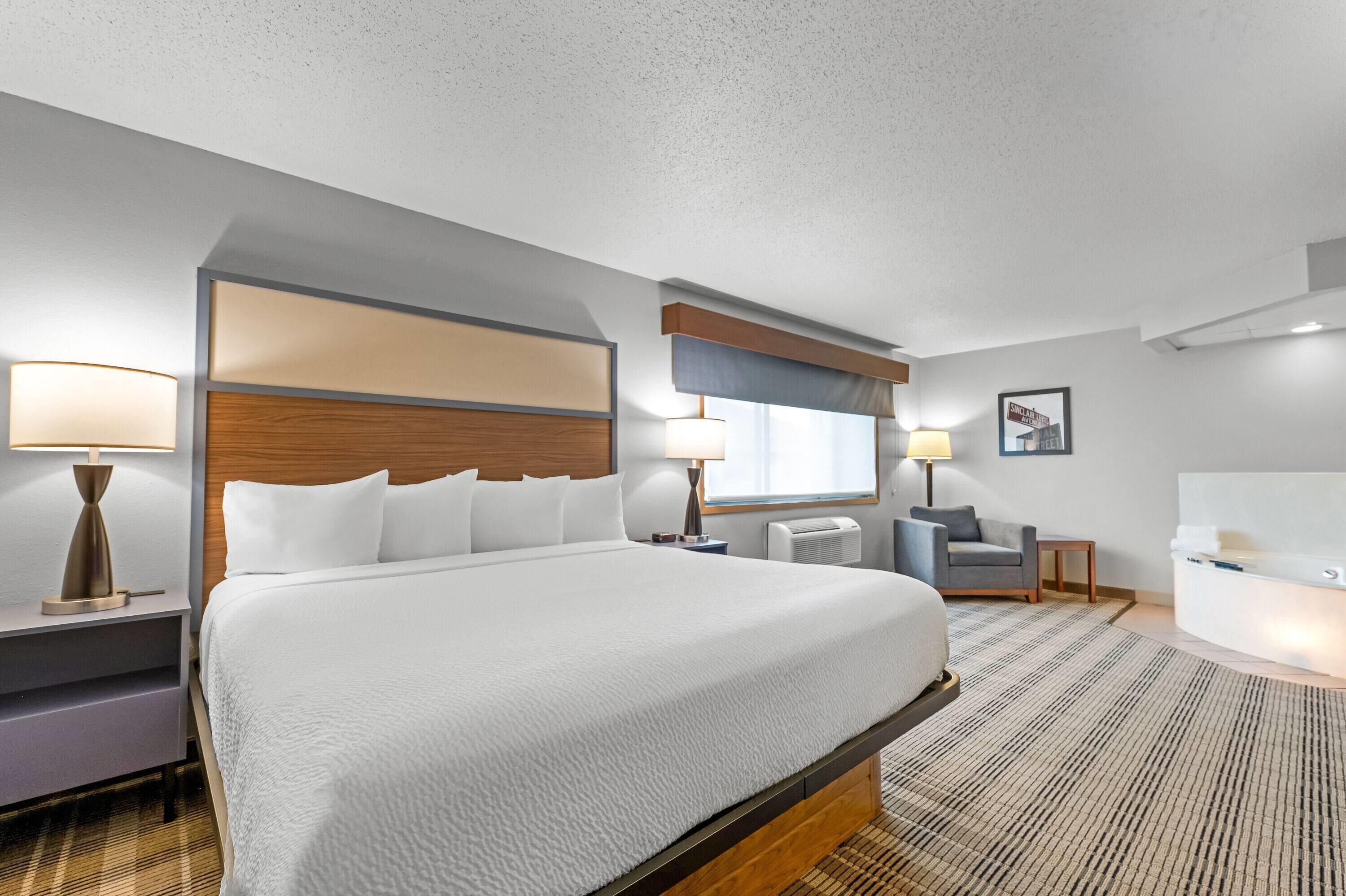 A hotel room with a large bed, bedside lamps, a small table, a chair, and a bathroom with a bathtub.