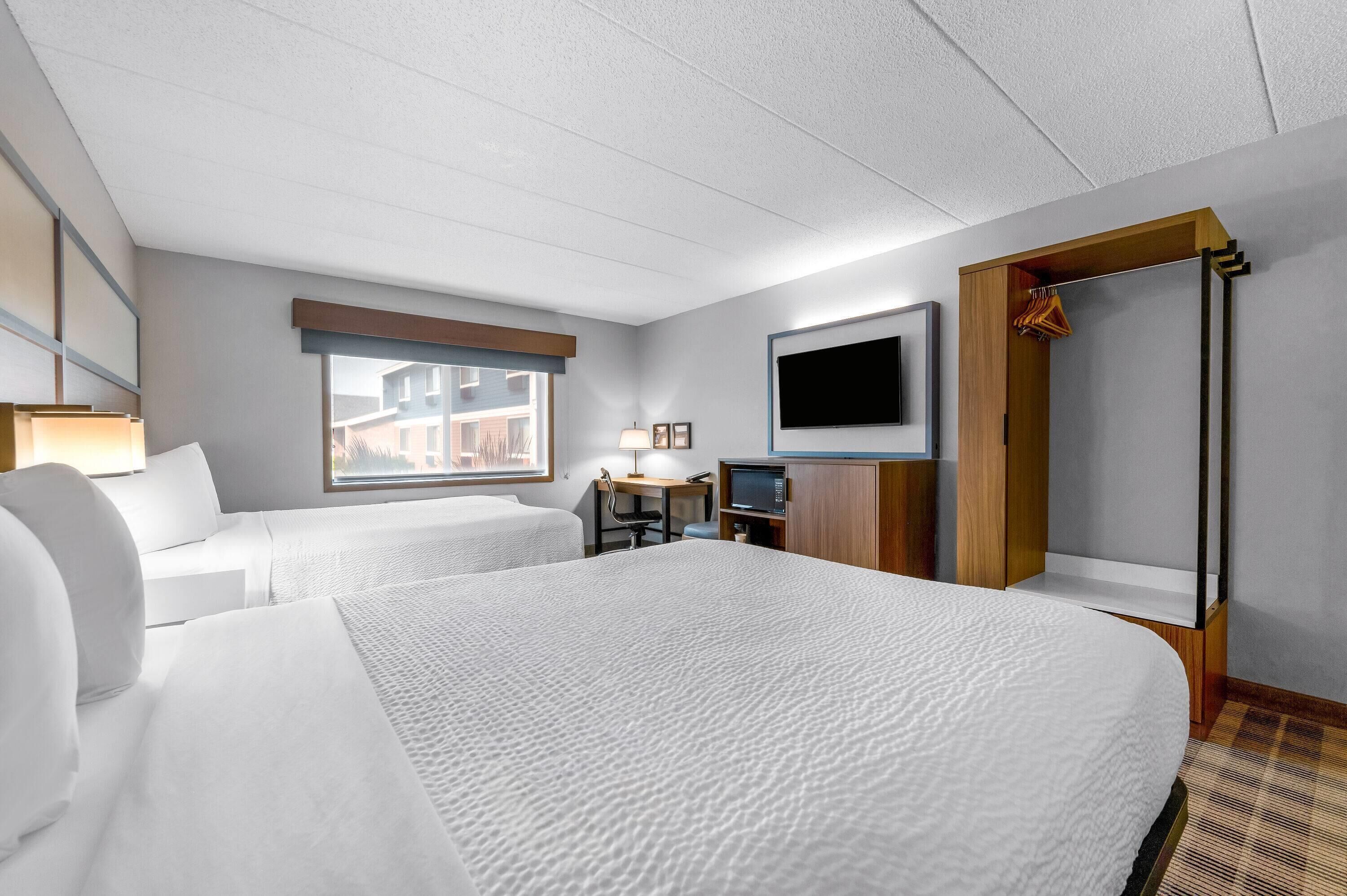 A hotel room with two beds, a desk with a computer, a TV mounted on the wall, and a window with a view of buildings.