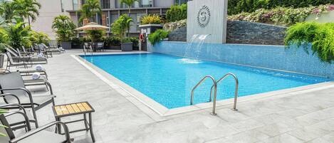 A swimming pool with lounge chairs and umbrellas, surrounded by greenery and a modern building.