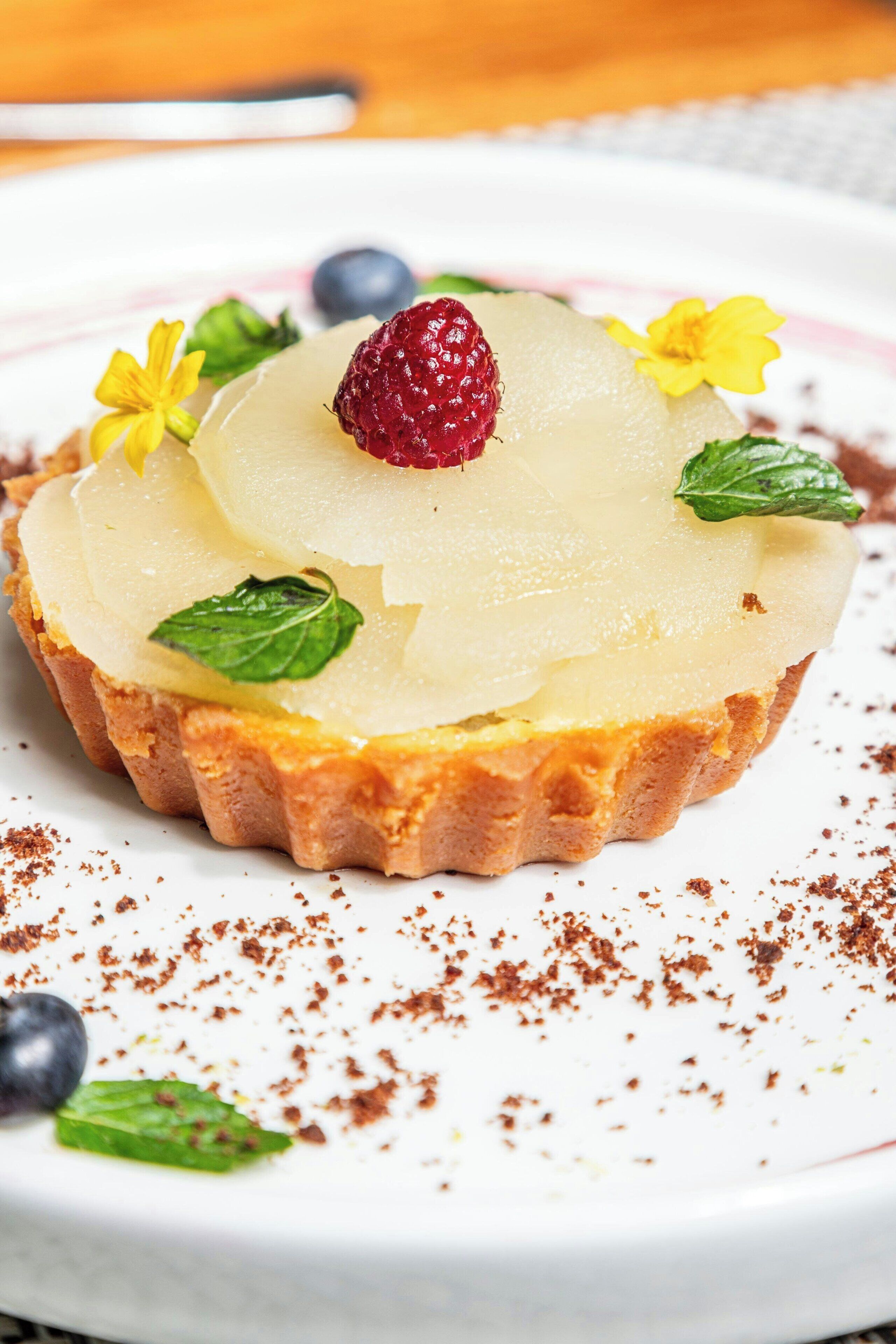 Ein Dessert mit säuerlichem Untergrund, bedeckt mit einer cremigen Schicht, einer Himbeere und verziert mit Minzblättern und gelben Blüten.