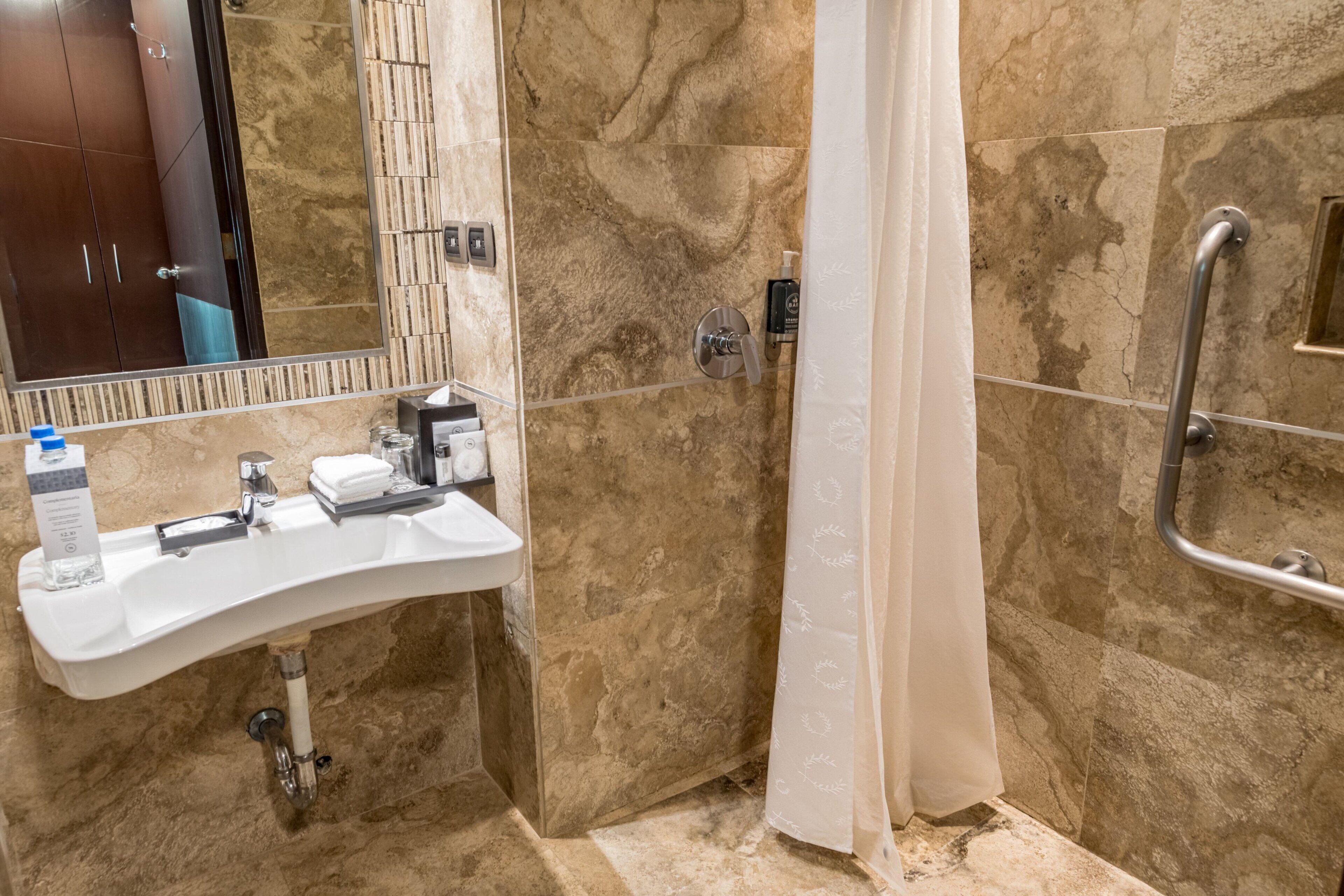 A bathroom with a sink, mirror, shower, and handrails.